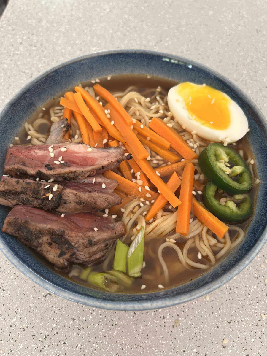 A bowl of Fiery Dragon Noodles topped with steak, carrots, jalapeños, and a soft-boiled egg, garnished with sesame seeds.