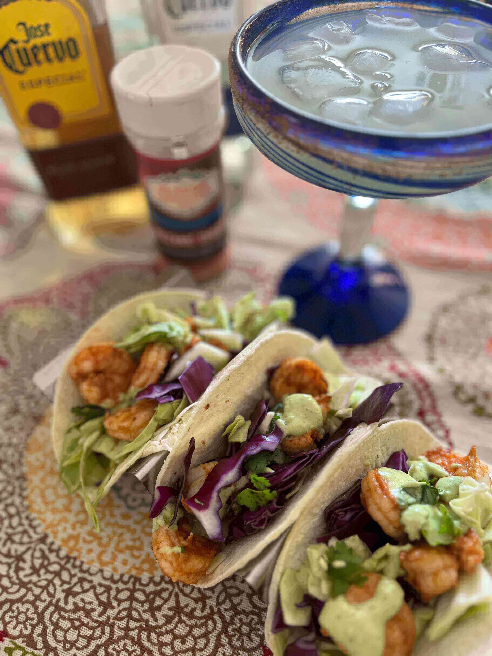 Volcanic Shrimp Tacos on a colorful table, garnished with lettuce, avocado sauce, and surrounded by drinks.