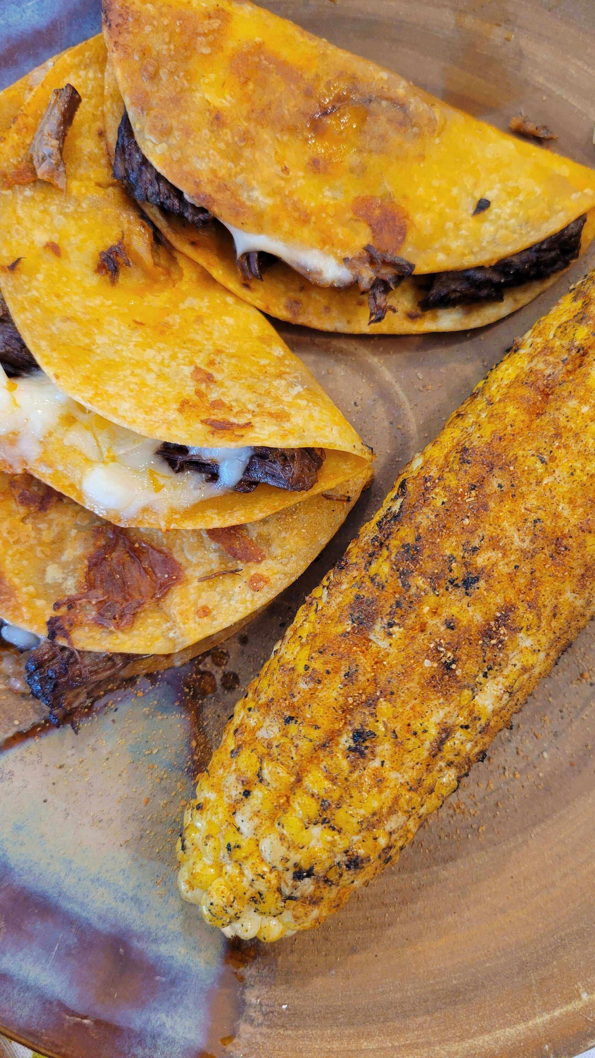 Plated Midnight Birria Tacos with melted cheese beside spicy grilled corn on the cob.
