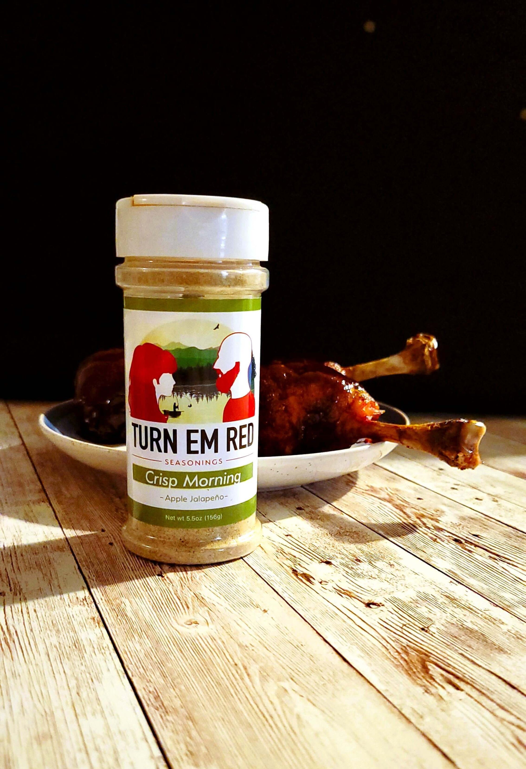 Seasoning jar of Turn Em Red Crisp Morning next to Apple Jalapeño BBQ Chicken Lollipops on a wooden table.