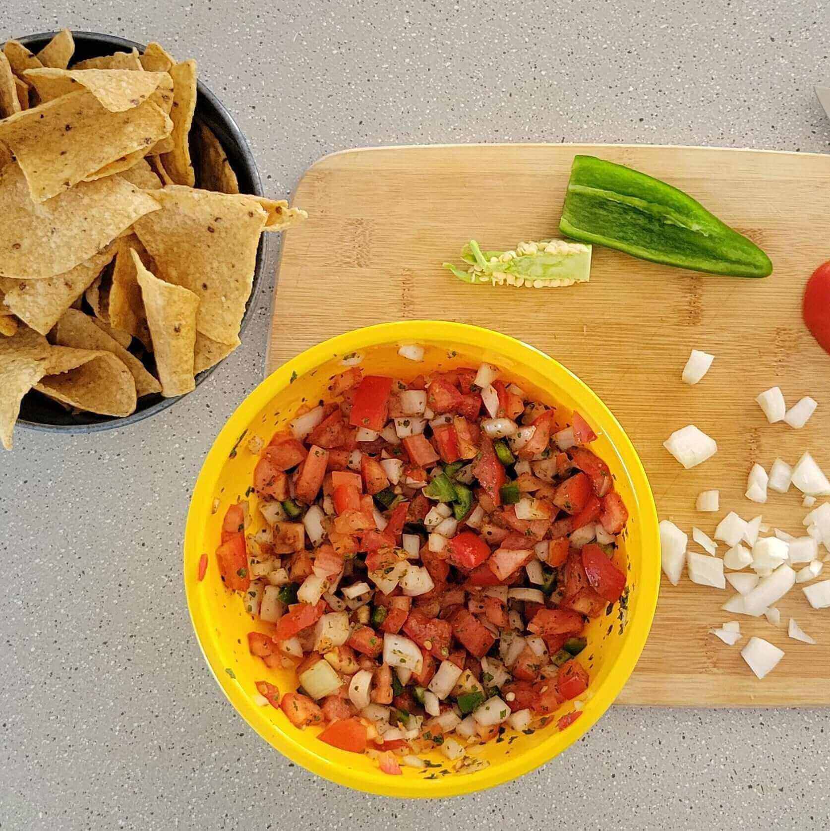 Howie’s No-Brakes Pico de Gallo in a yellow bowl with chips, a green pepper, and chopped onions on a wooden cutting board.