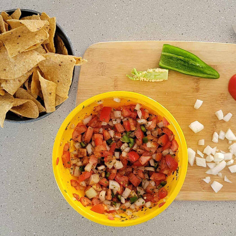 Howie’s No-Brakes Pico de Gallo in a yellow bowl with chips, a green pepper, and chopped onions on a wooden cutting board.