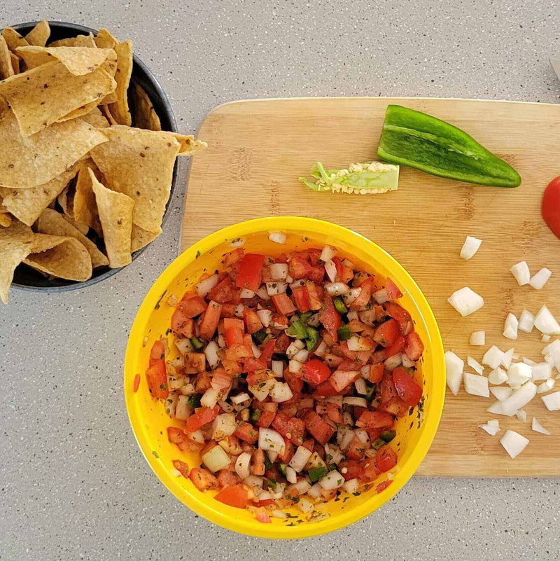Howie’s No-Brakes Pico de Gallo in a yellow bowl with chips, a green pepper, and chopped onions on a wooden cutting board.