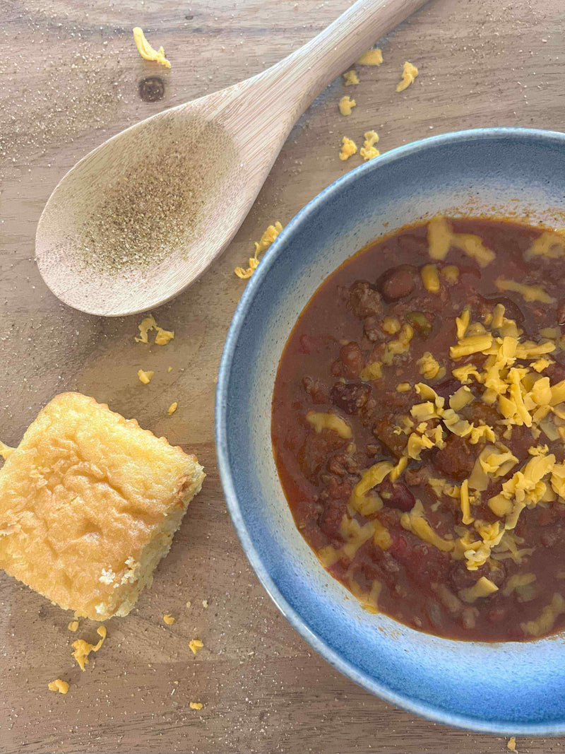 A delicious bowl of "Chuck It in the Pot" Western Dawn Crockpot Chili topped with cheese, served with cornbread and a wooden spoon.