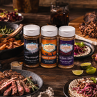 Three jars of Flavor Expedition seasoning on a wooden table with food in the background