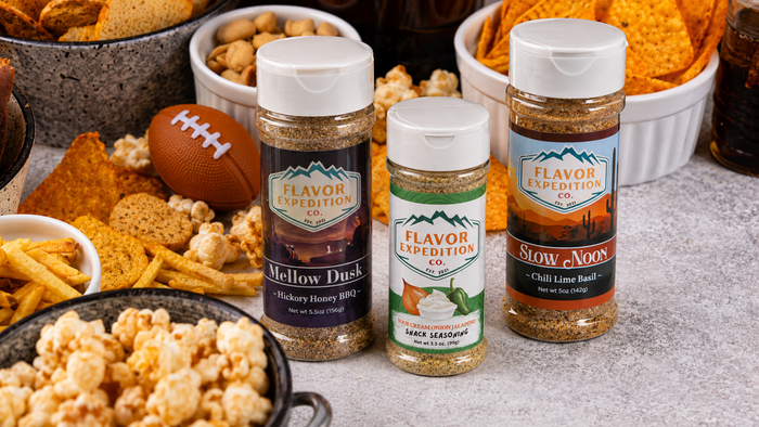 Three Flavor Expedition Co. seasoning bottles displayed on a gray stone surface surrounded by snack foods like chips, popcorn, crackers, peanuts, and a football.