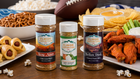 Three Flavor Expedition Co. seasoning bottles displayed on a wooden table surrounded by snacks and a football, with pretzels, hot dogs, chips, celery, wings, popcorn, and dips.