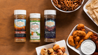 Three Flavor Creation Co. seasoning bottles arranged on a wooden tabletop with snacks around them, including pretzels, tortilla chips, chicken nuggets, and a dipping sauce.