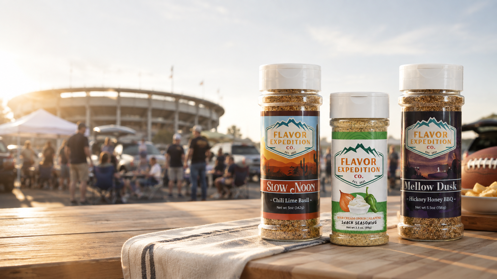 Three Flavor Expedition Co. seasoning bottles displayed on a wooden picnic table outdoors at a football game, with a stadium and crowd blurred in the background.