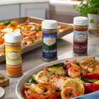 Three bottles of Flavor Expedition seasoning on a kitchen counter with cooked shrimp and vegetables.