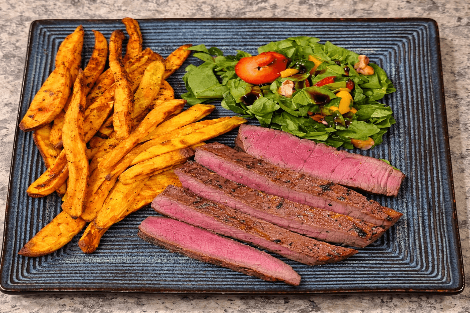 Plated dish with sliced steak, sweet potato fries, and a salad on a textured black plate.