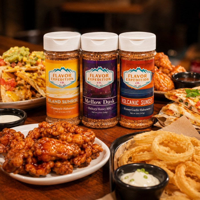 Three bottles of Flavor Expedition spices on a table with food in the background