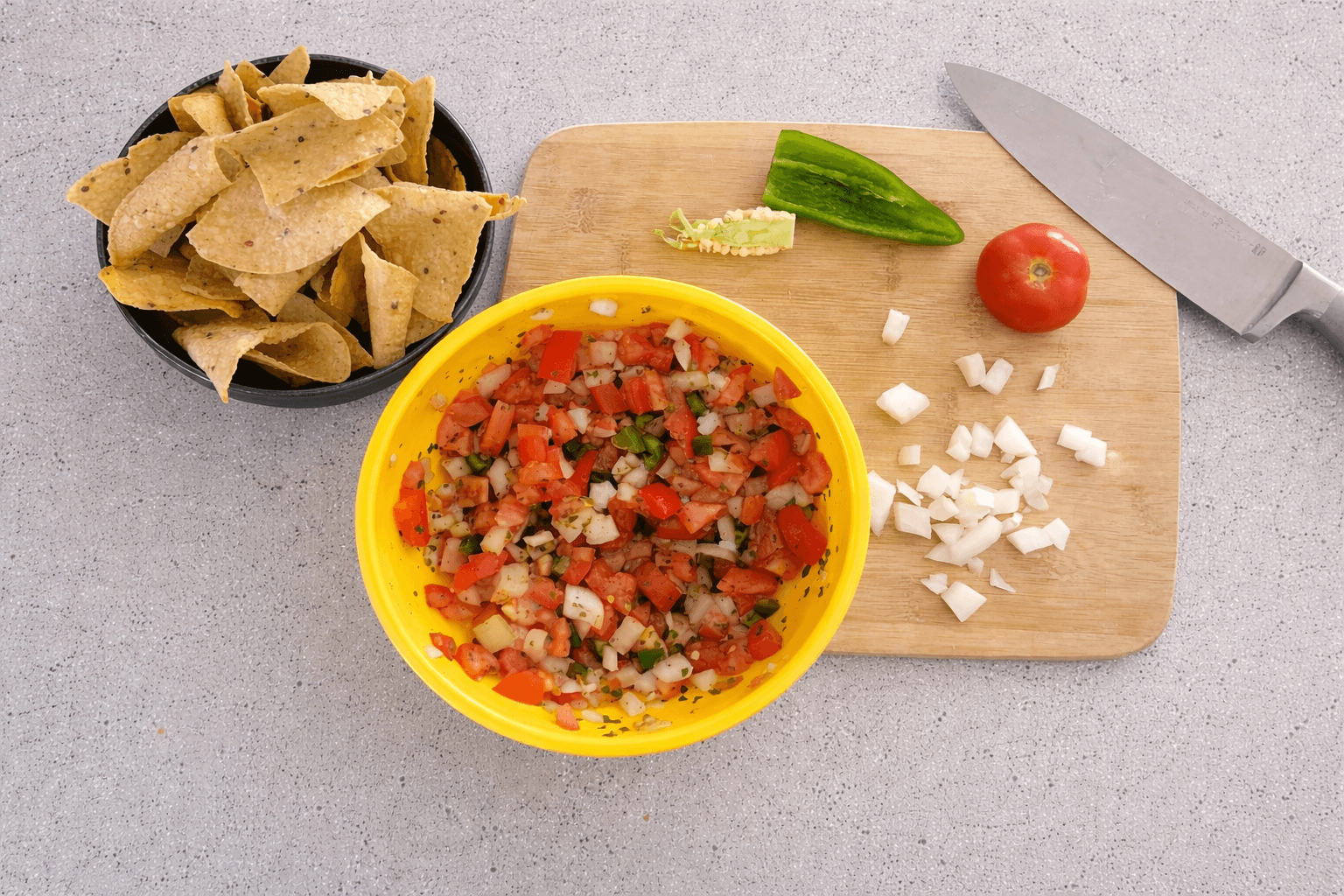 Yellow bowl of salsa with tortilla chips, tomato, and jalapeno on a cutting board.