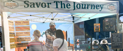 People interacting under a 'Savor The Journey' tent at an outdoor event.