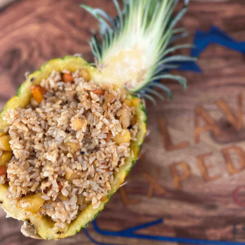 A stuffed pineapple bowl filled with brown rice and small orange fruit pieces, placed on a wooden board with engraved text.