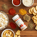 Spices, popcorn, crackers, and chips on a wooden surface with a jar of seasoning.