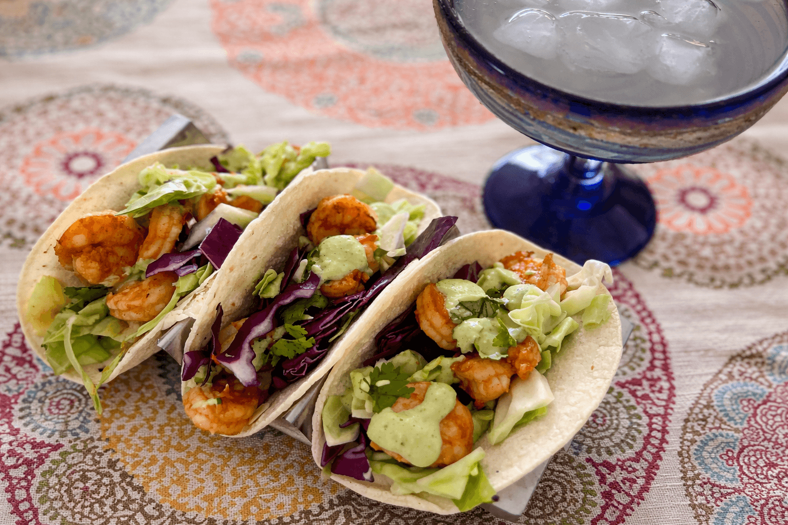 Three shrimp tacos with guacamole on a patterned tablecloth with a cocktail in the background.