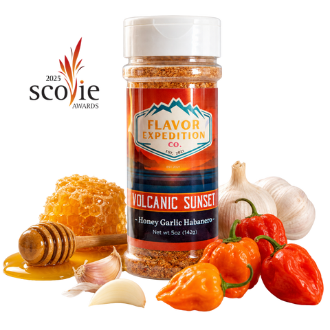 A bottle of Flavor Expedition Co. 'Volcanic Sunset' seasoning stands on a white background surrounded by food props including honeycomb, honey, a honey dipper, garlic cloves, red and orange habanero peppers, and chili peppers. [pack-1]